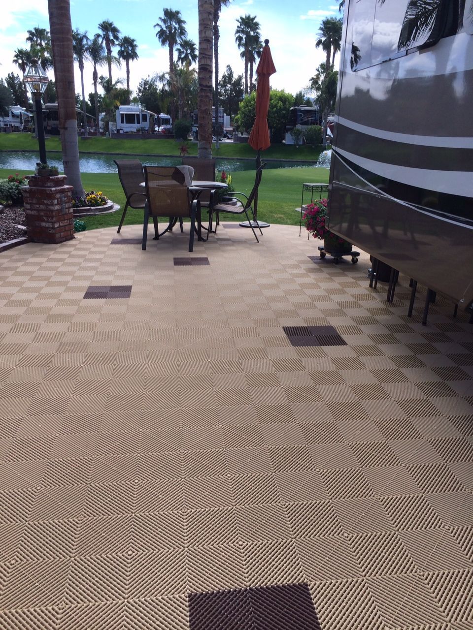 RV patio with checkered tan and brown tiles, table, chairs, umbrella, and a view of palm trees and a lake.