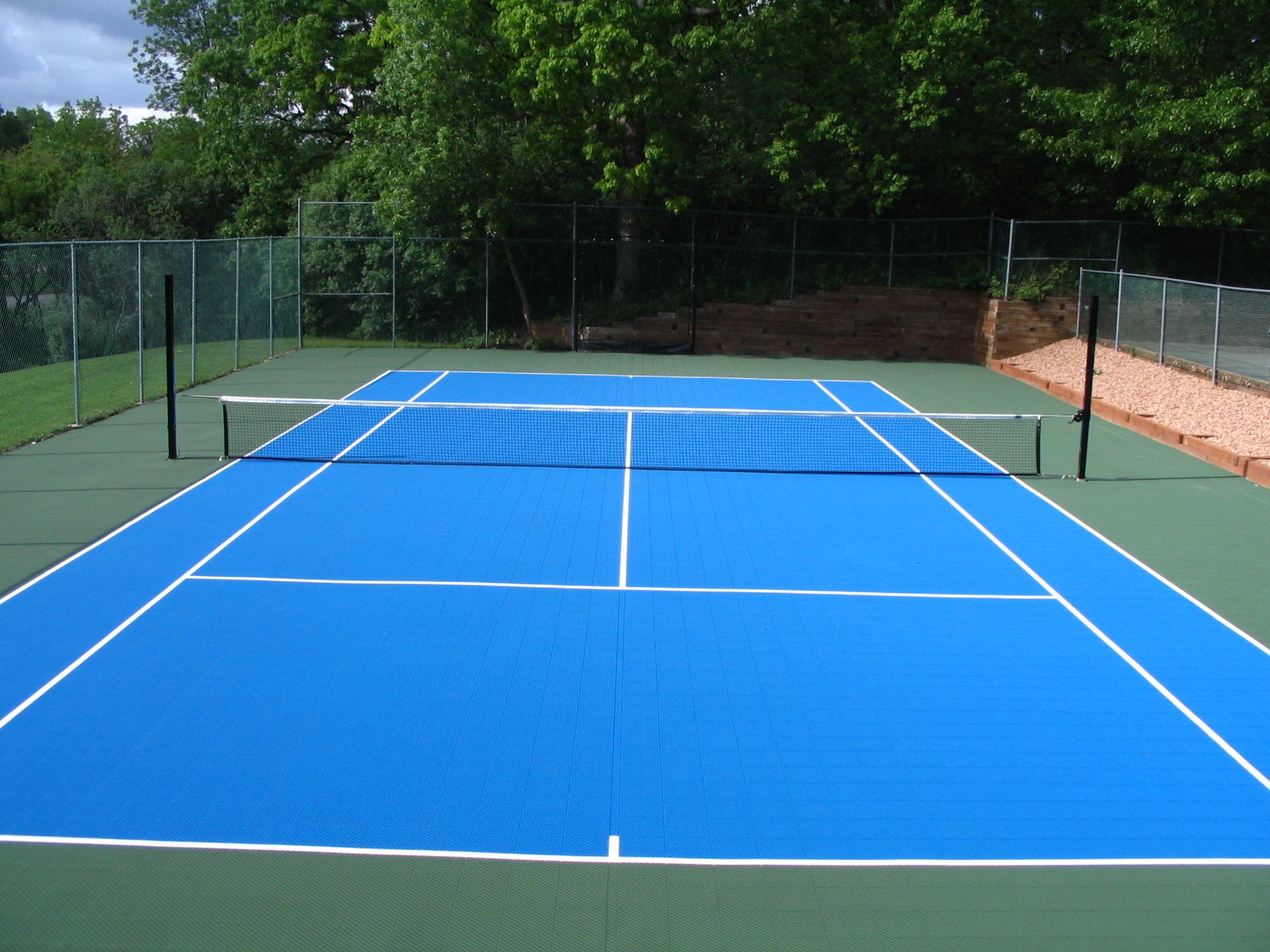 Blue and green tennis court with white lines and net, surrounded by a fence and trees.