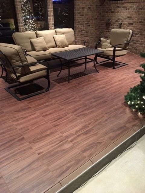 Outdoor seating area with wood-look tile floor, beige sofa, chairs, and black table against a brick wall.