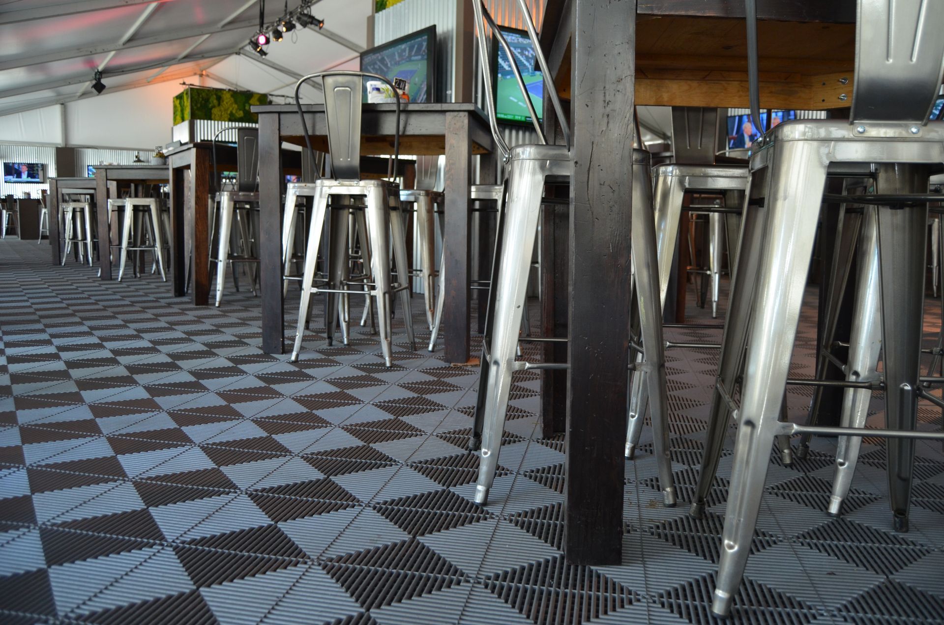 Rows of metal bar stools and tables on a checkered gray and blue floor under a tent.