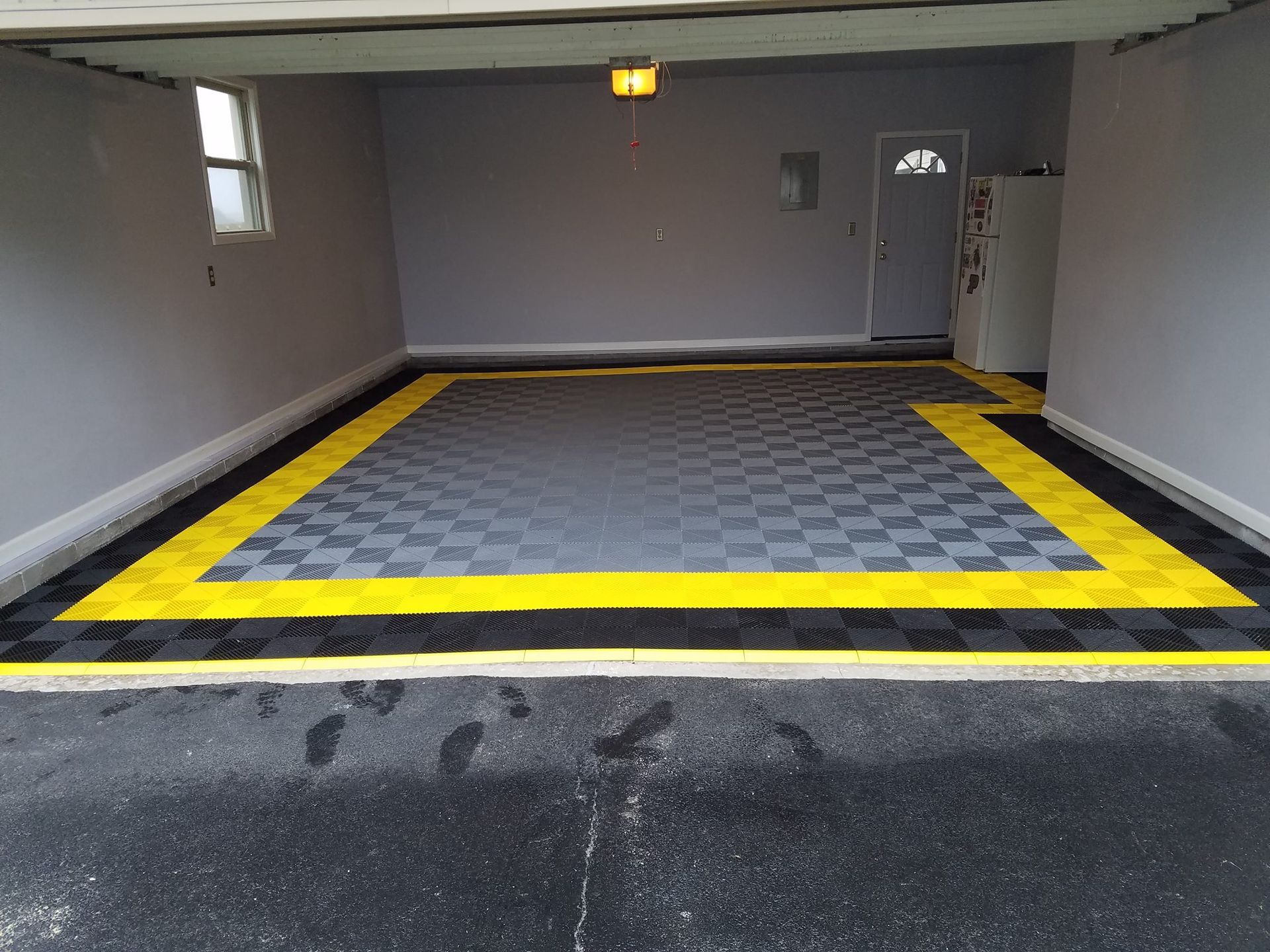 Garage interior with gray and yellow checkered flooring. Paved driveway leads to the garage entrance. Gray walls and a small window are visible.
