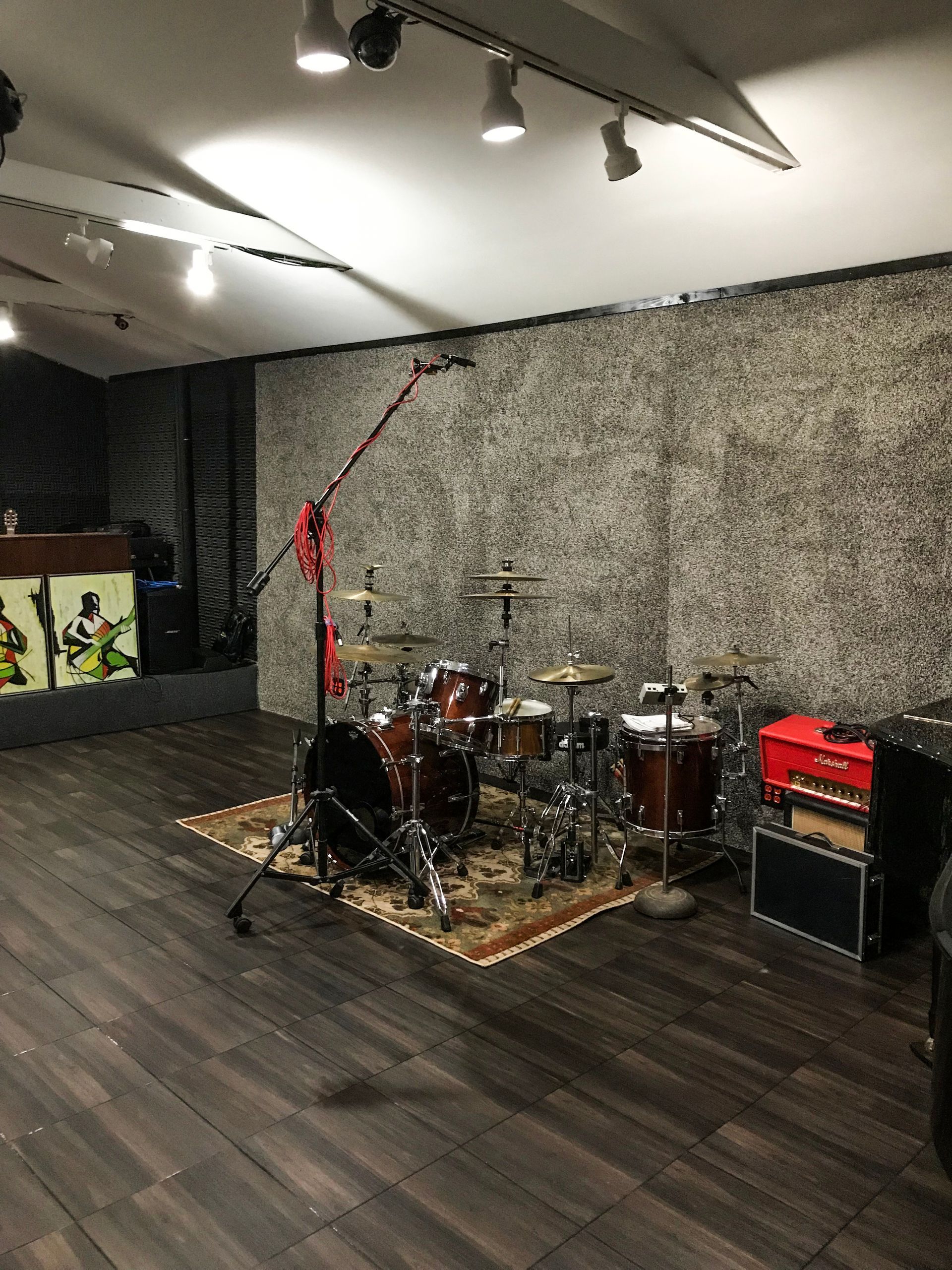 Drum set and amplifiers in a music studio, featuring a textured wall, overhead lighting, and dark wood flooring.