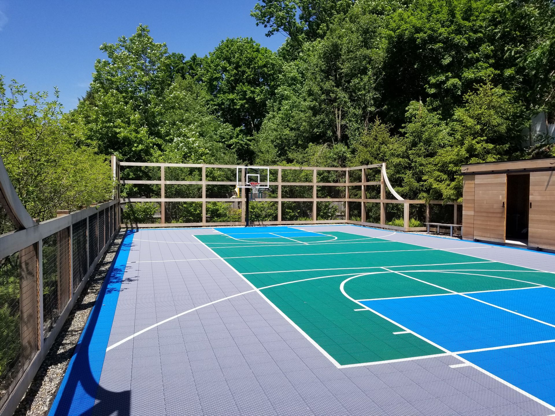 Basketball court with blue, green, and gray tiles. Wooden fence and trees surround the court.
