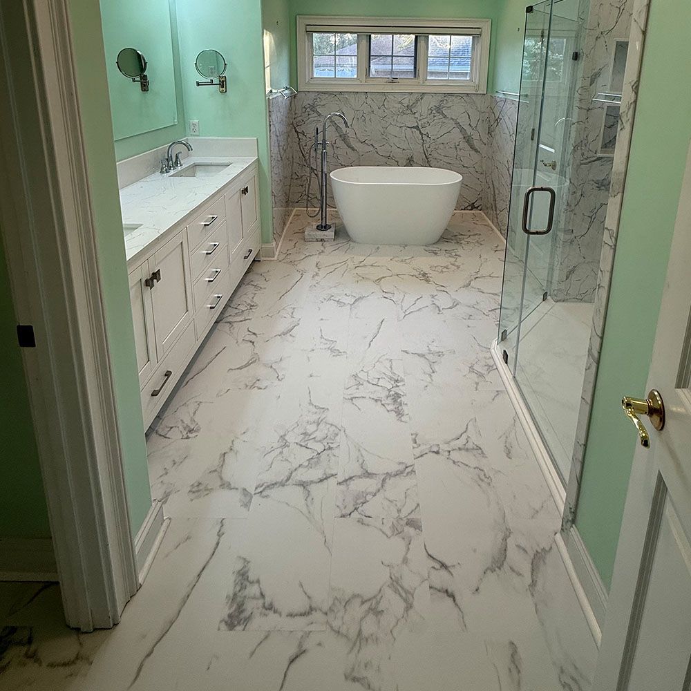 Spacious bathroom with marble-look flooring, a white soaking tub, and a glass shower. Light green walls and white cabinets.