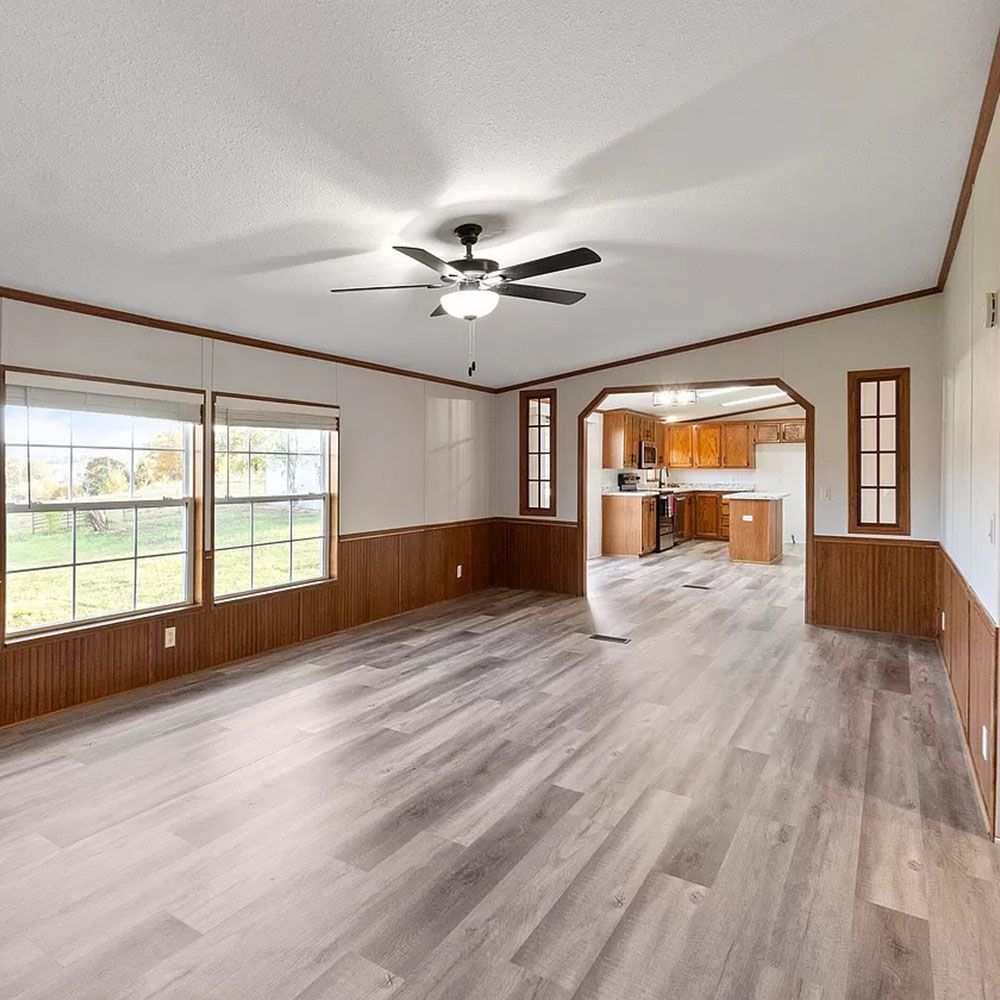 Empty living room with wood trim, gray flooring, and view into the kitchen.