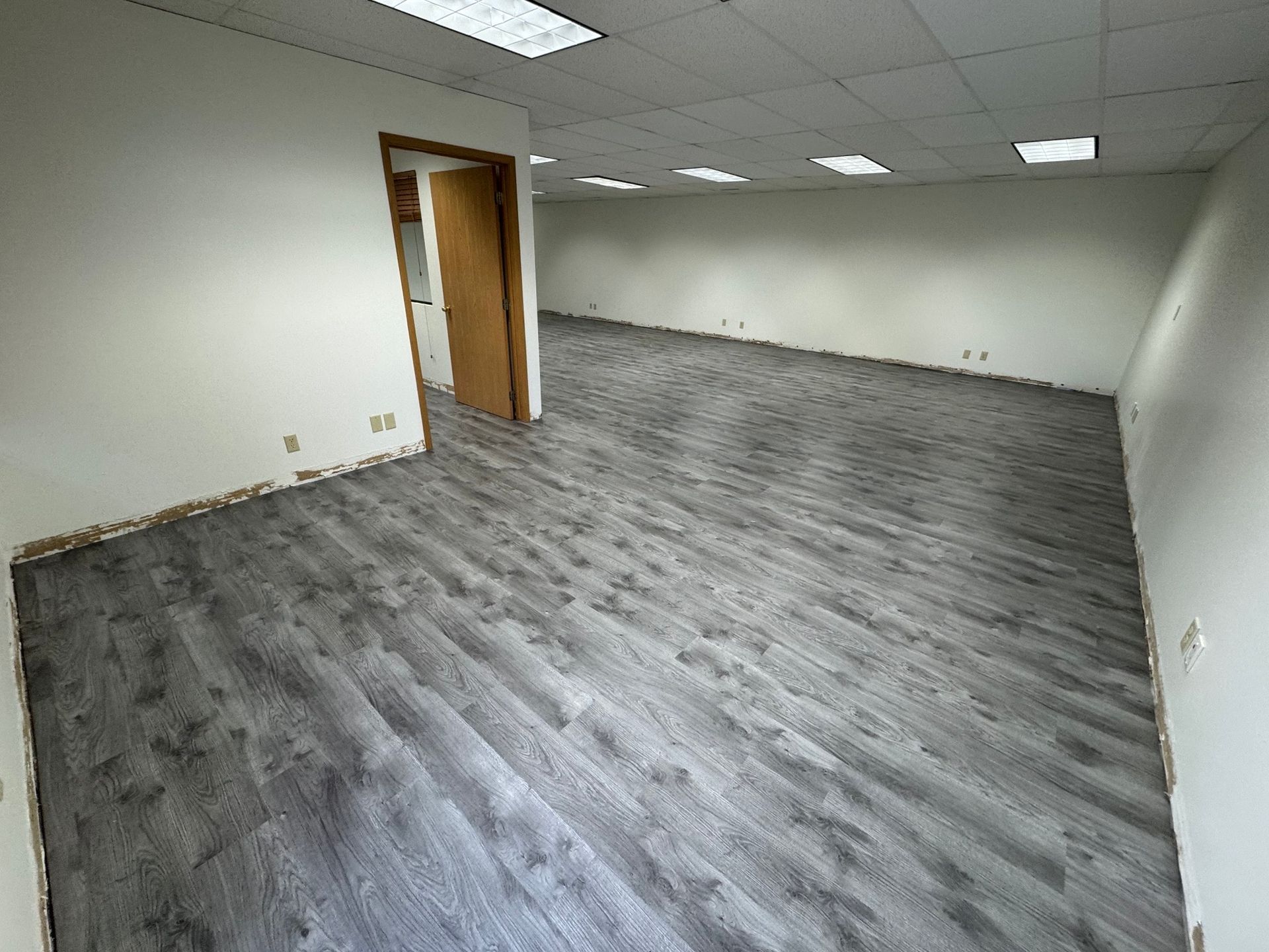 empty room with grey wood-looking flooring and white walls, under fluorescent lights, door open