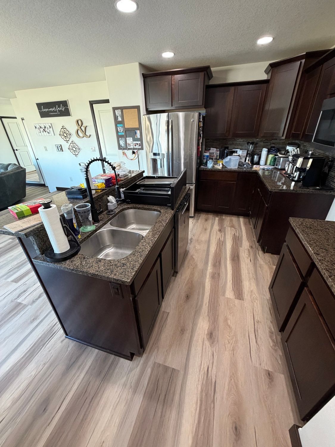 Modern kitchen with dark cabinets, stainless fridge, island sink, and wood-look floor.