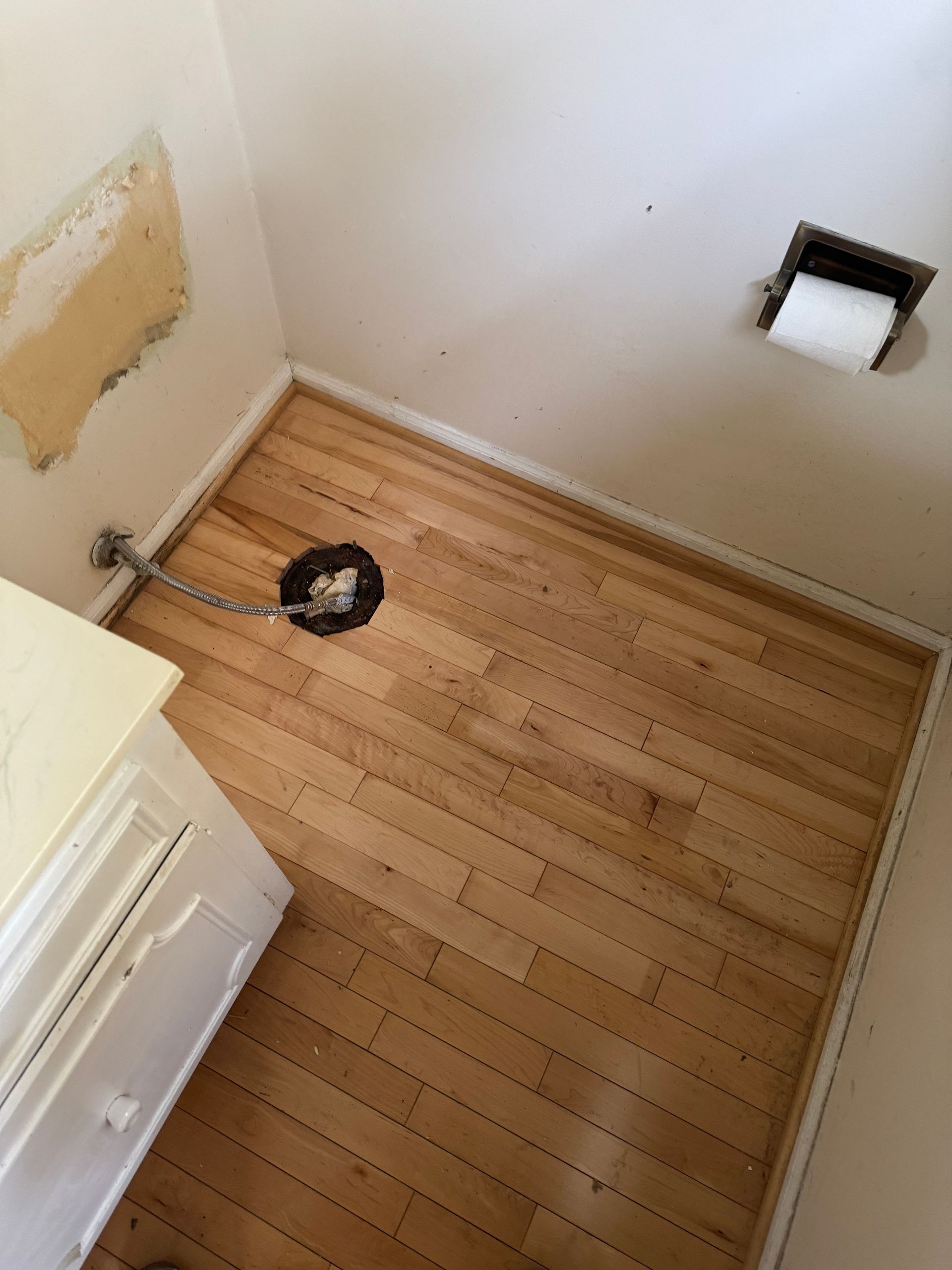 Small empty bathroom with wood floor, a toilet paper holder, and exposed wall patching near the corner.
