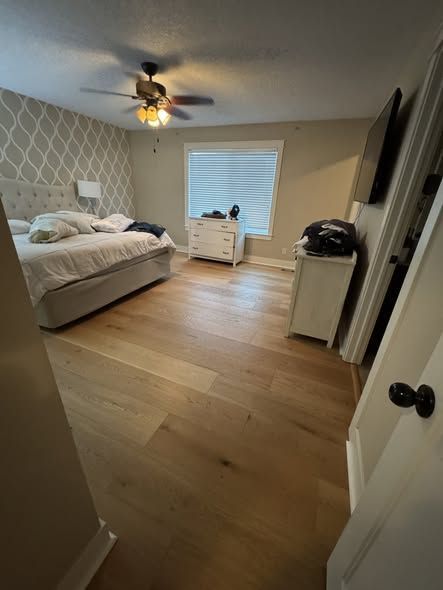 Bright bedroom with a bed, ceiling fan, white dresser, wall-mounted TV, and wood floors