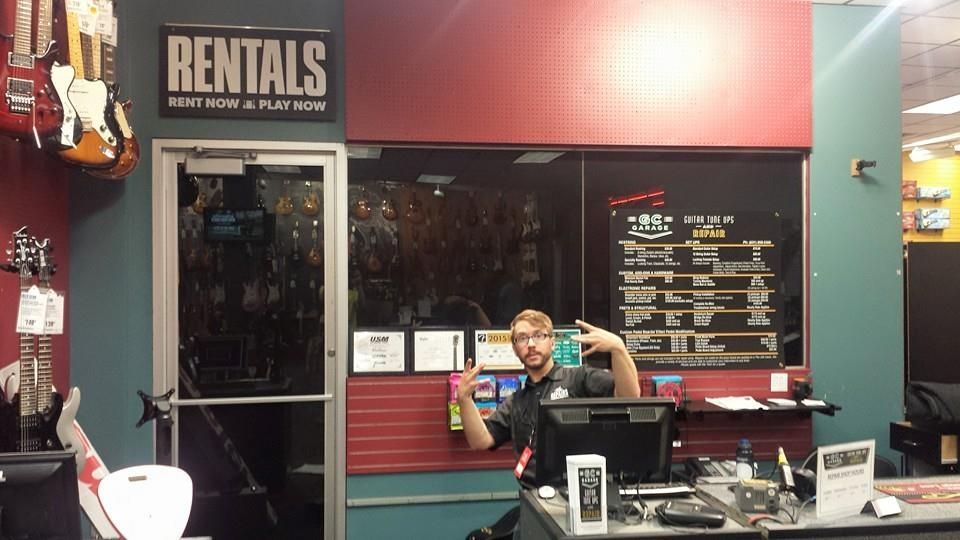 A person at a music store rental desk makes a rock-and-roll hand gesture while sitting in front of guitars and signs.
