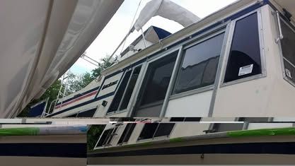 A side-angle view of a white, multi-level houseboat with sliding glass doors, docked in a natural, tree-lined setting.