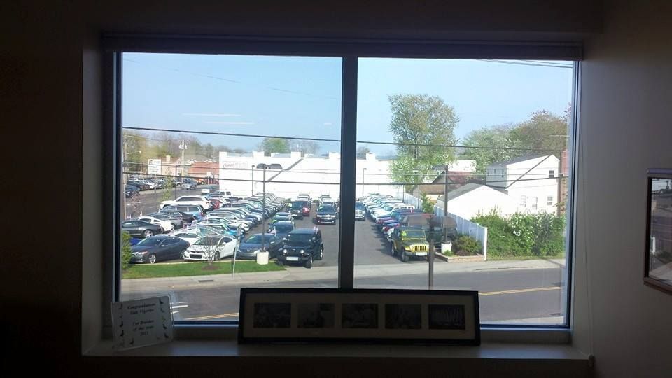 A view through a large window overlooking a parking lot filled with cars and a white building under a clear blue sky.