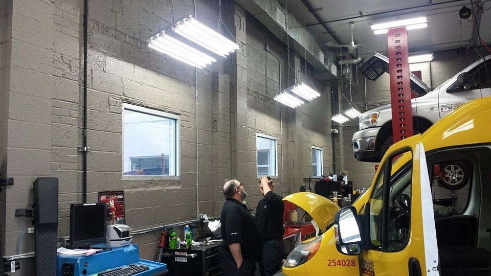 Two technicians talk in a repair shop next to a raised white vehicle and a yellow van with its hood open.