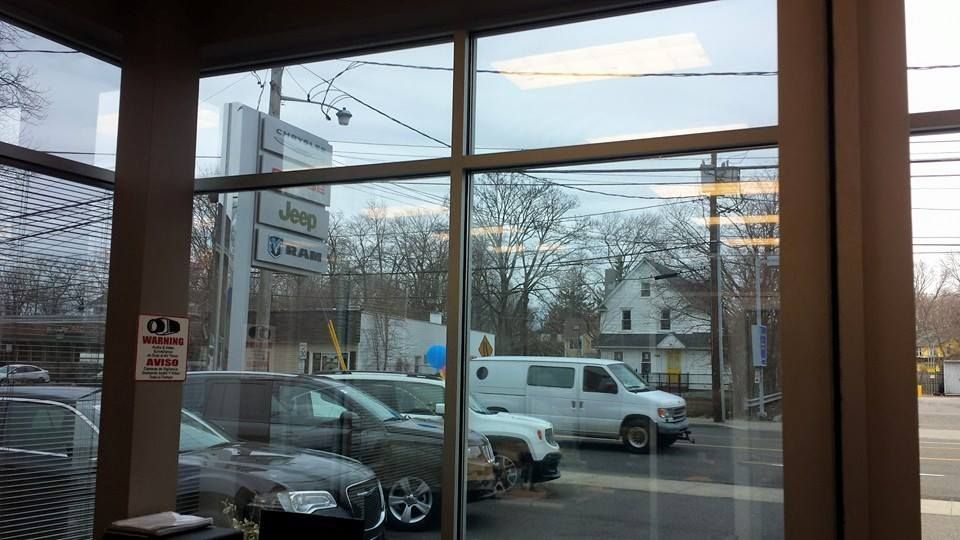 View from a window showing a car dealership sign, parked vehicles, a white van on the road, and a house in the distance.