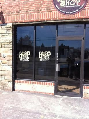 The storefront of The Hop Shop, a home brew supply store featuring brick walls and glass windows with branded decals.