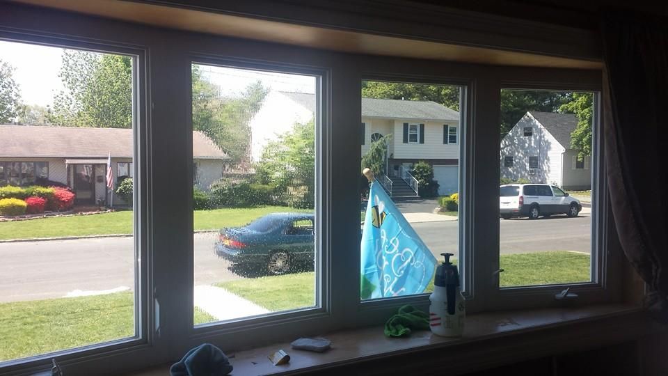 A view from inside looking through a four-pane bay window at a suburban neighborhood street with houses, cars, and a flag.