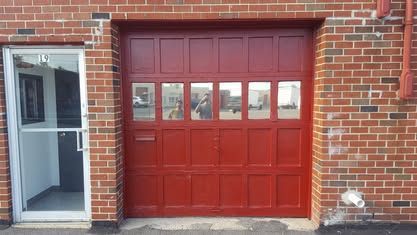Red garage door with rectangular windows next to a glass door in a brick wall.
