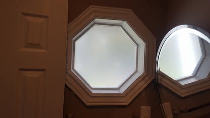 An octagonal, white-framed window on a beige wall, with a partial reflection of the same window in a nearby mirror.