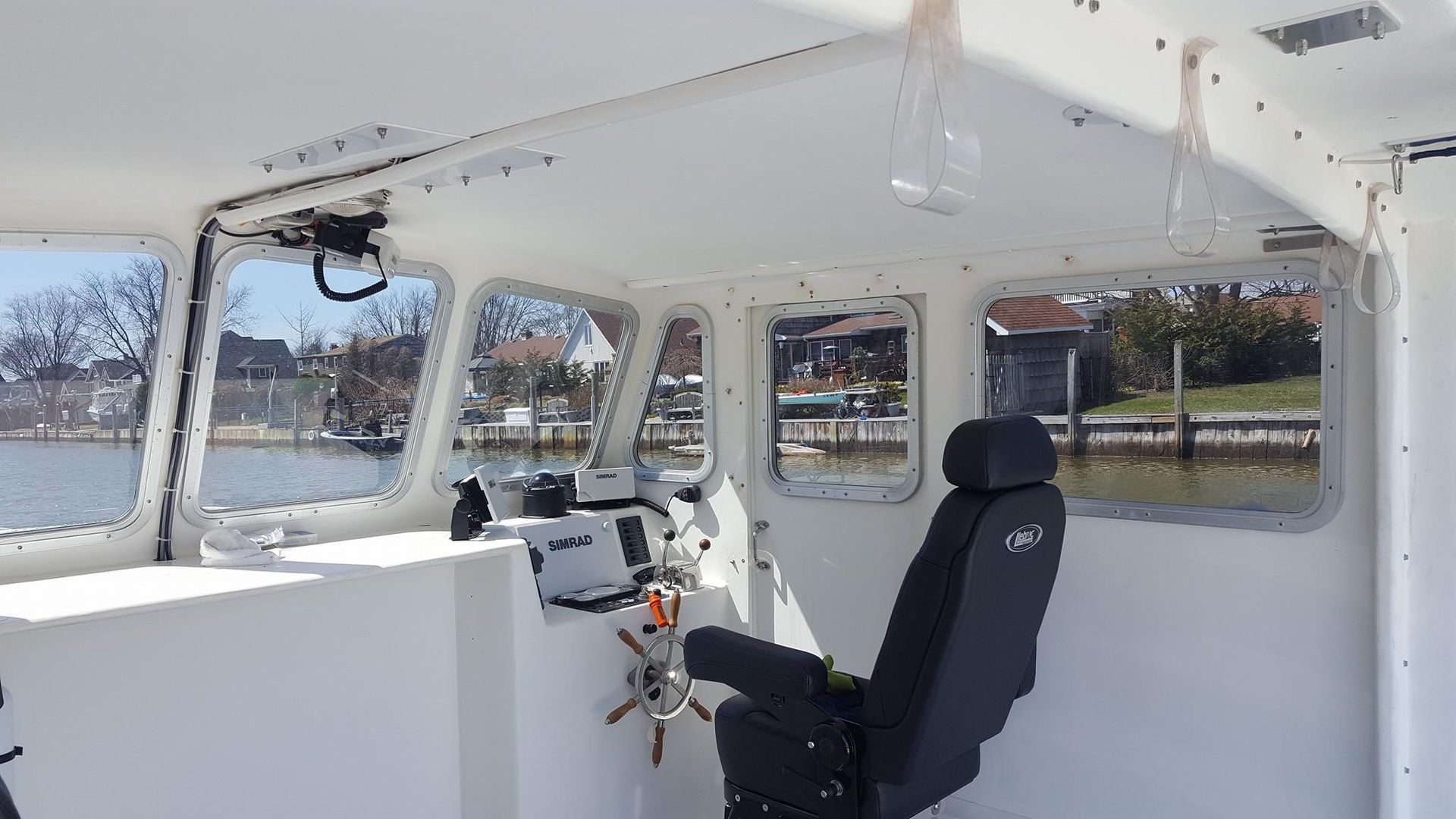 The interior of a boat cockpit featuring a black pilot seat, steering wheel, and navigation console facing water views.