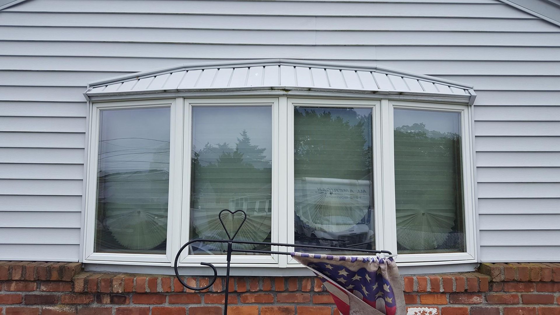 A bow window with a metal roof on a house with light grey siding and a brick base, partially obscured by a garden flag.