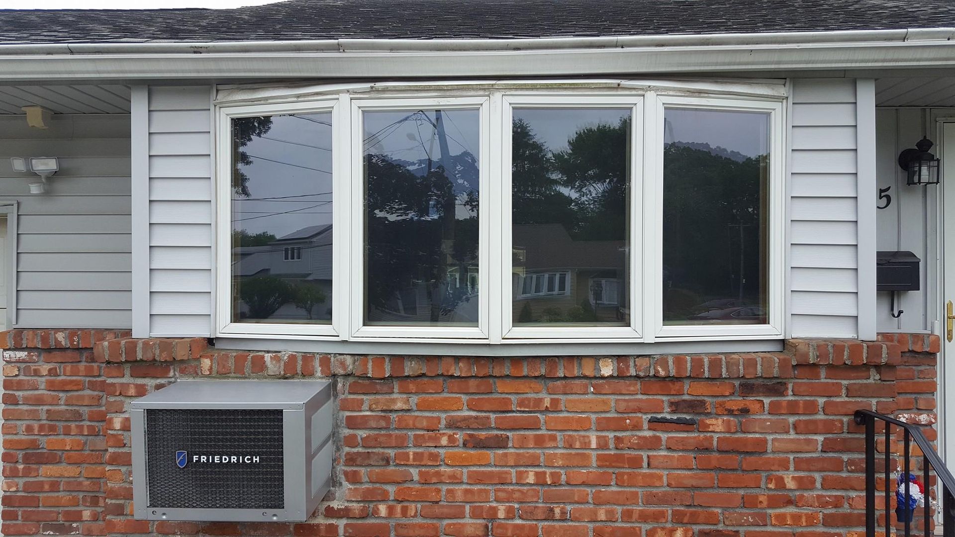 A four-pane bay window on a red brick house with light gray horizontal siding and a window air conditioning unit.