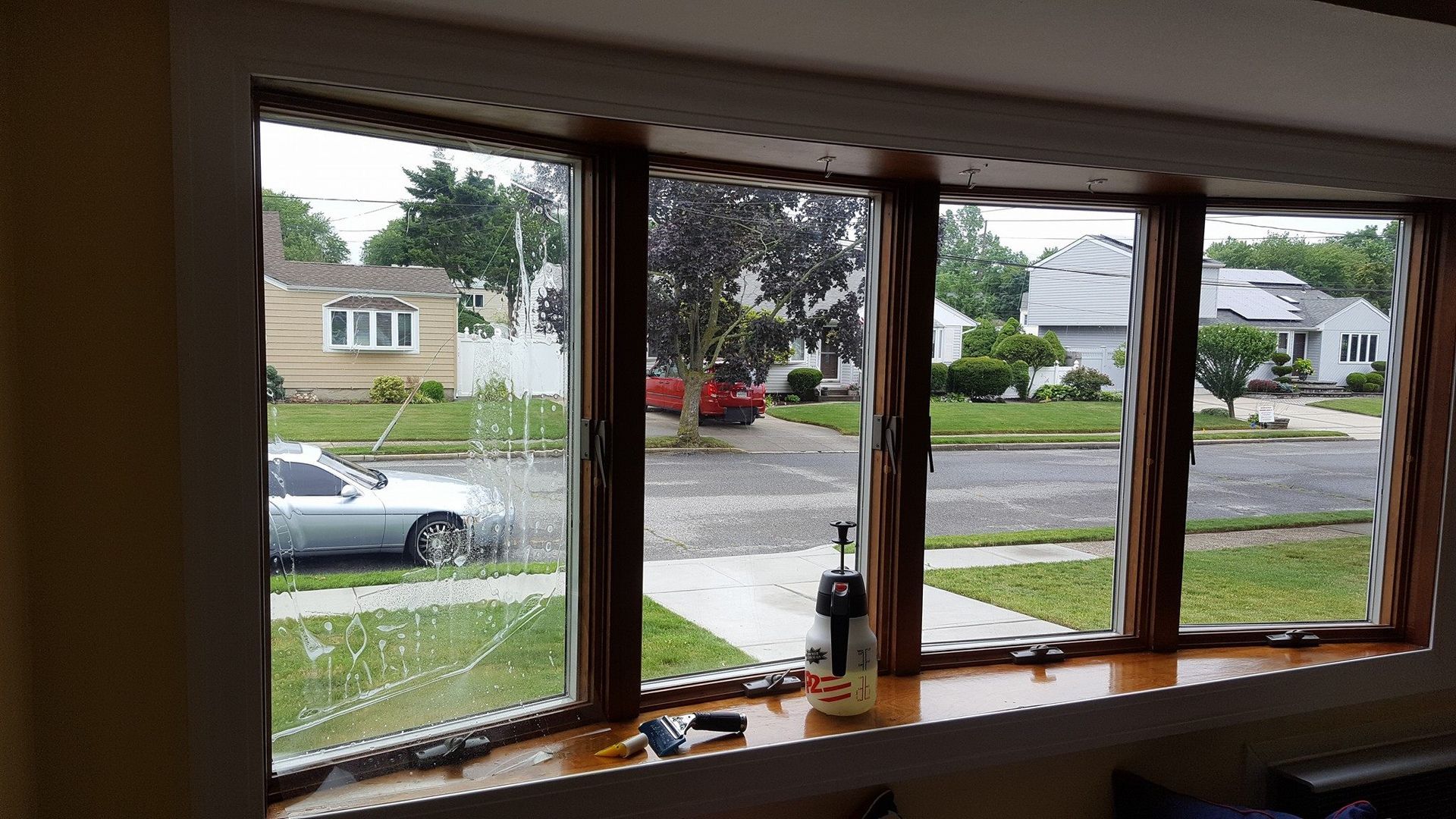 A view from inside looking out through a multi-pane bay window at a residential street, with cleaning spray on the sill.