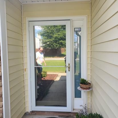 A white storm door on a house with light yellow siding, reflecting a person standing outside on a sunny day.