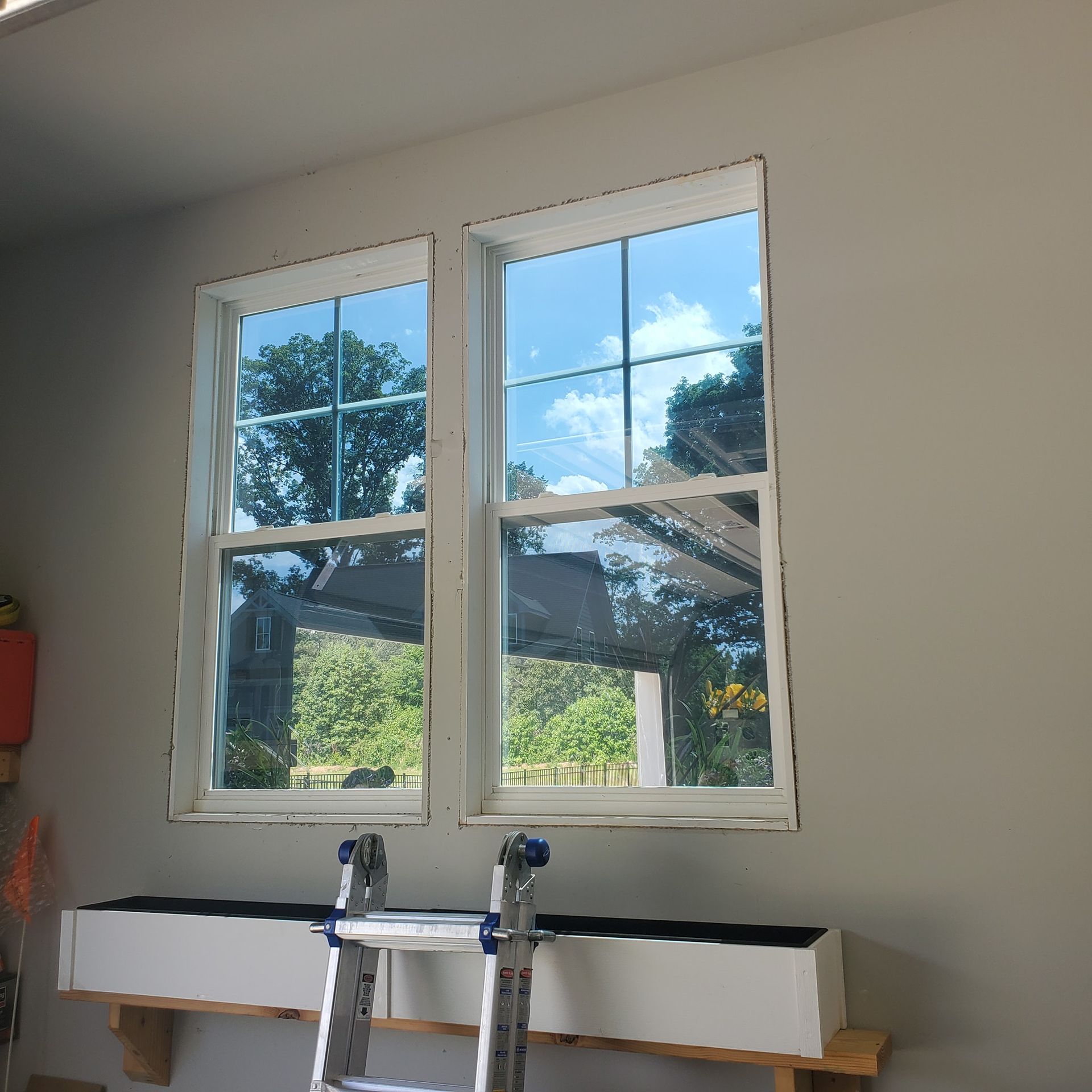 Two white-framed windows with grid panes set in a light grey wall above a white shelf with a silver ladder leaning on it.