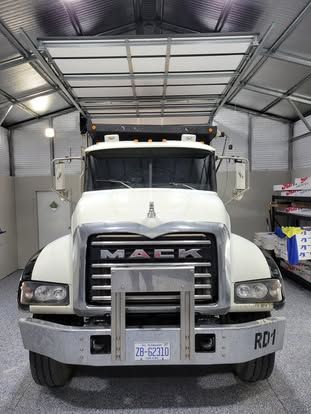 A white Mack dump truck parked inside a bright, clean garage with metal walls and a shiny, speckled concrete floor.