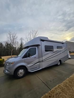 A silver Mercedes-Benz Sprinter motorhome parked on a concrete driveway during a cloudy day.
