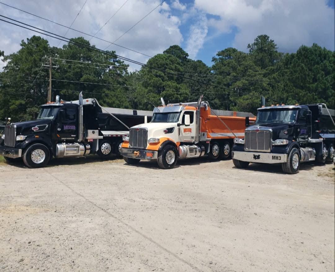 Three dump trucks parked side-by-side on a gravel lot; two are black, and the middle truck is white and orange.