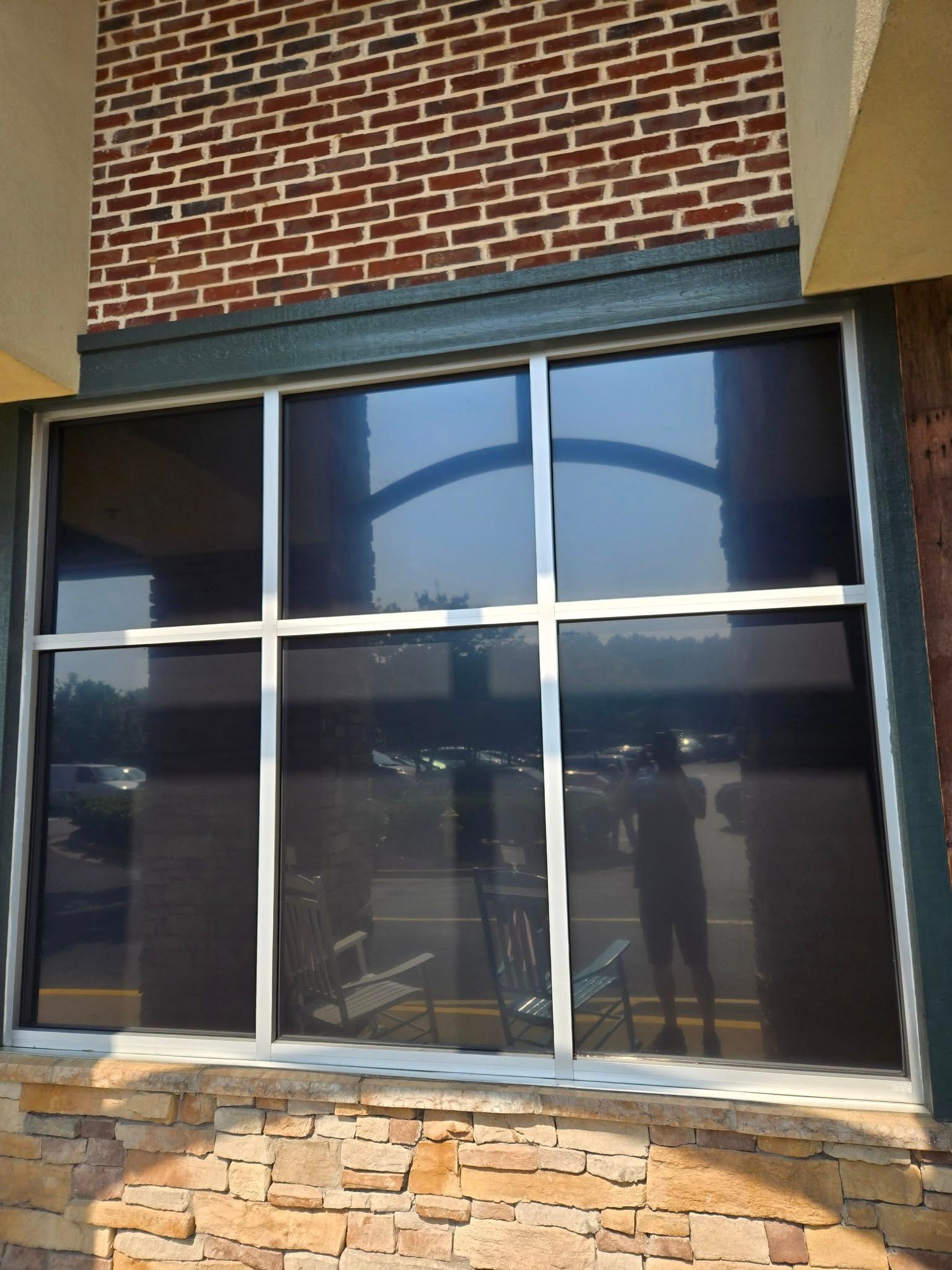 A large glass window with a grid frame reflects a parking lot, trees, and a person standing inside.