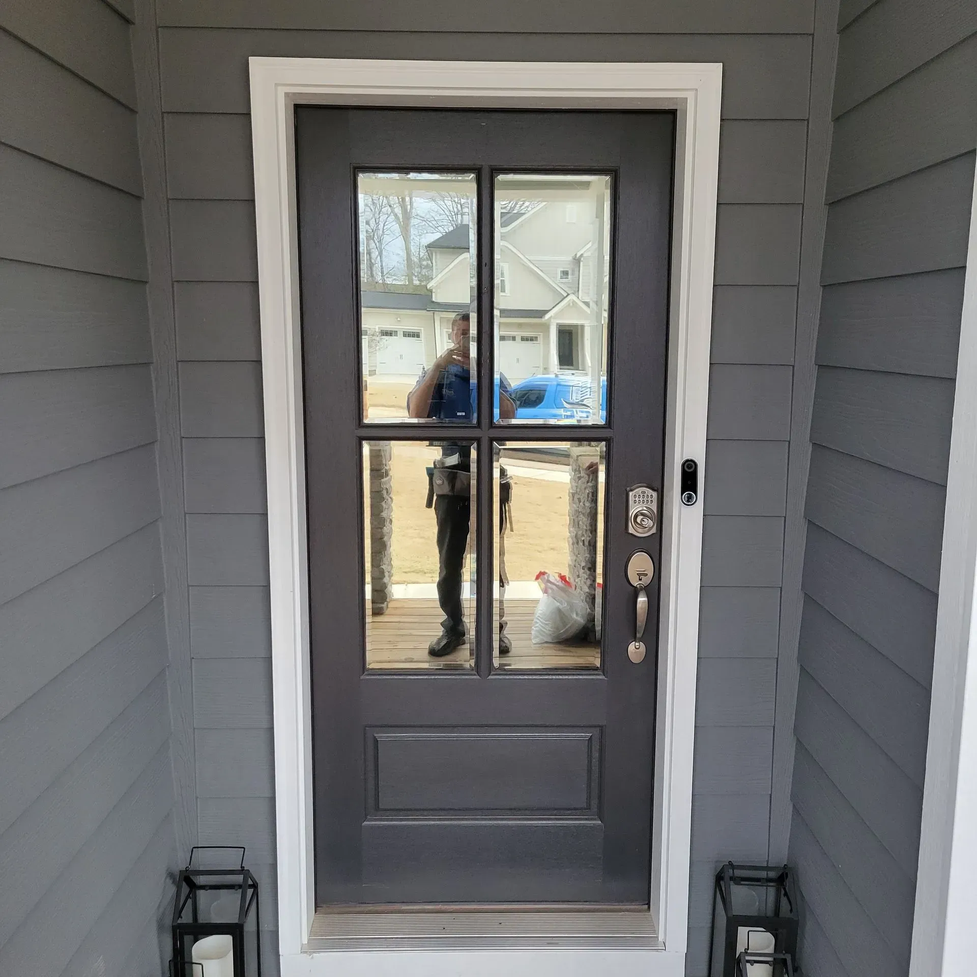 A dark gray front door with four glass panes set in a white frame, flanked by gray siding and two lanterns on the porch.
