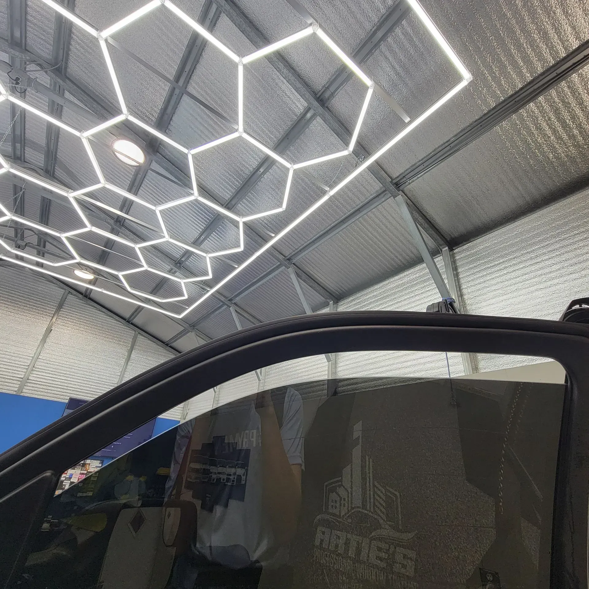 A close-up view through an open car window showing a hexagonal LED ceiling light grid mounted on a reflective wall.