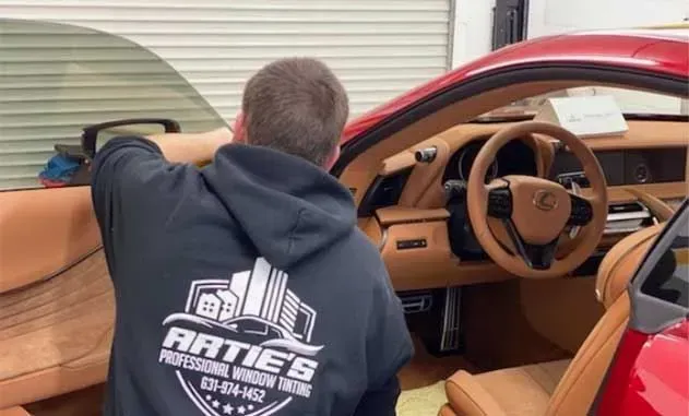 A professional window tinter in a branded hoodie applies tint to the driver-side window of a red car with a tan interior.