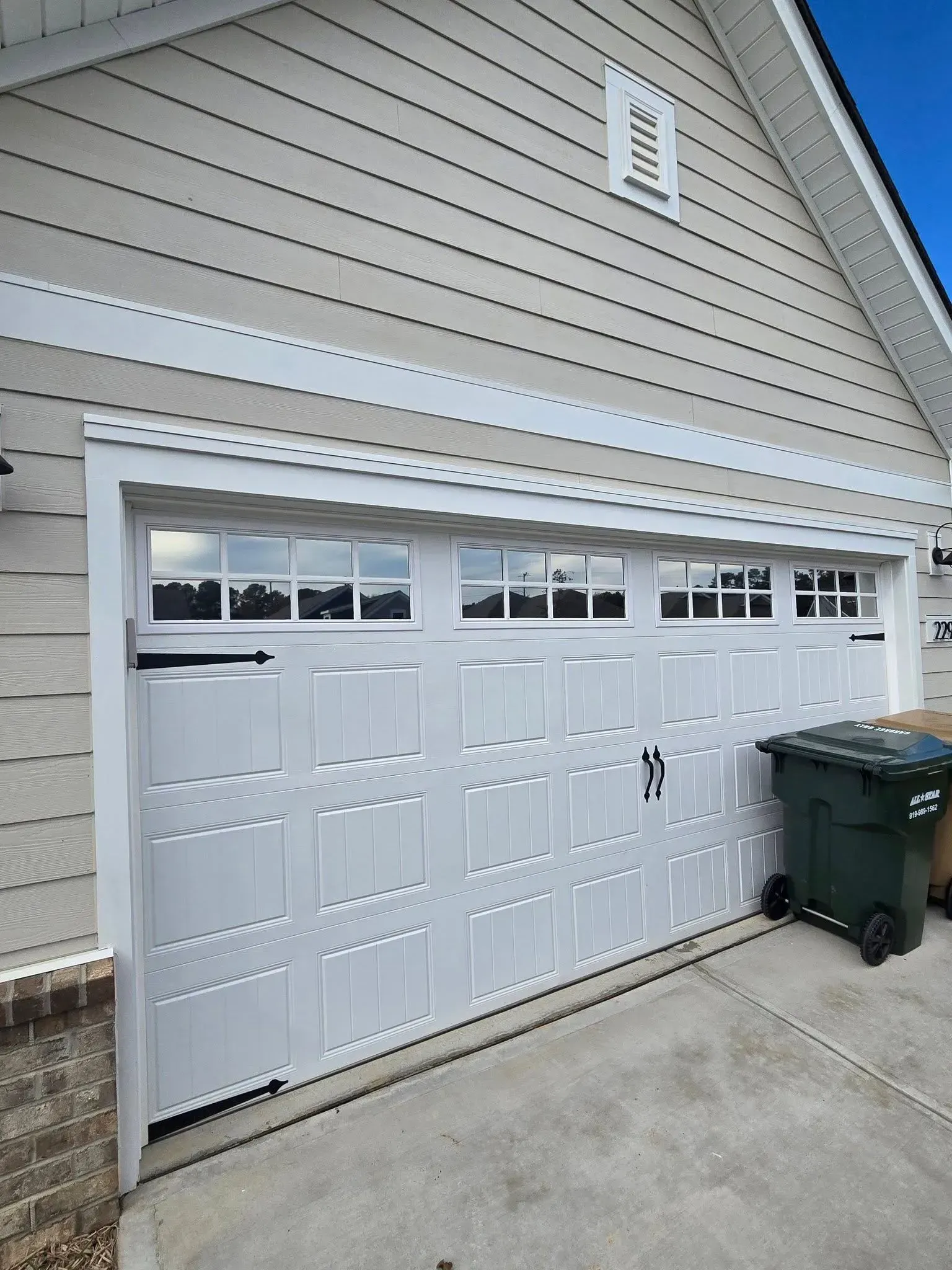 A white residential garage door with rectangular windows and decorative black hardware, next to a dark green trash bin.