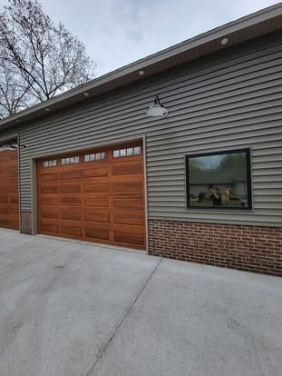 A detached garage with gray horizontal siding, brick base, brown wooden doors, a black-framed window, and a wall lamp.