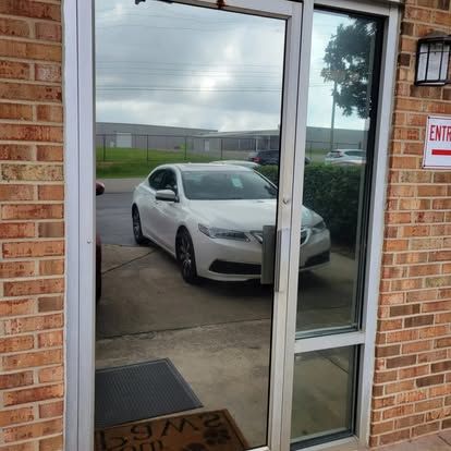 A glass door with a metal frame reflects a white sedan parked in a paved lot in front of a brick building.