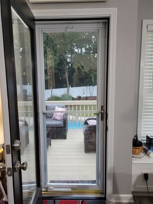 A view through an open dark-framed door to a deck with outdoor furniture, a small pool, and trees in the background.