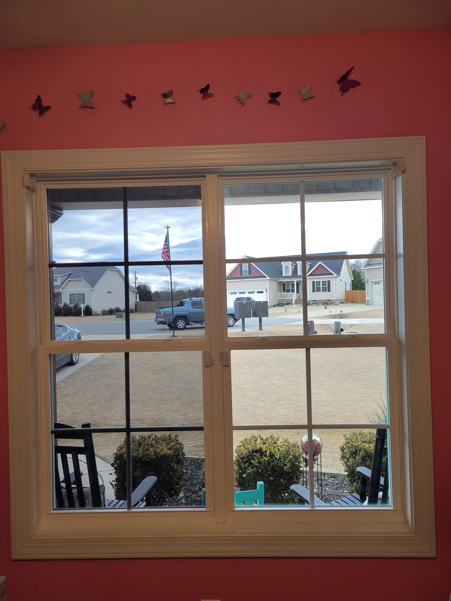 A window looks out onto a suburban street with houses and a parked truck, with butterfly decorations on the red wall above.