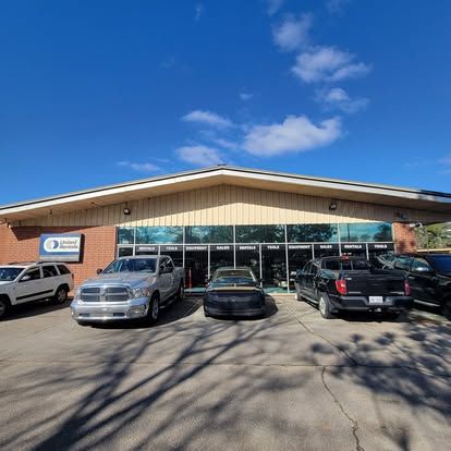 A one-story brick and tan building with a wide glass front, featuring parked vehicles out front under a blue sky.