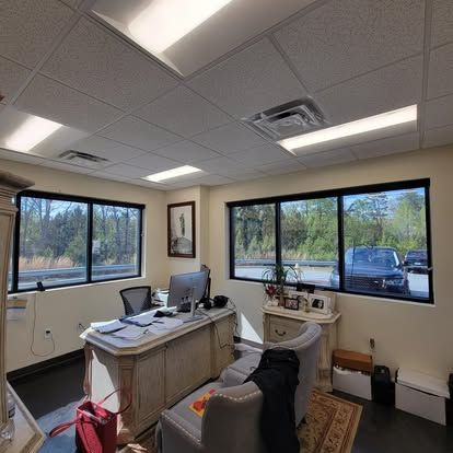 A modern, well-lit office with a desk, chair, framed art, and large windows overlooking a parking area and trees.