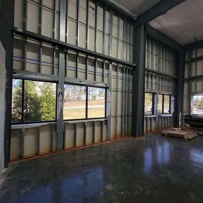 Construction site interior with steel wall framing and large window panels above a dark, polished concrete floor.