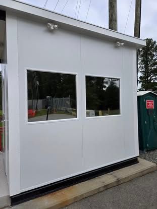 A white, modular security booth with two large windows and external lighting, situated outdoors next to a portable toilet.