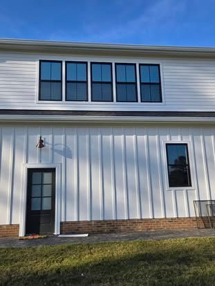 White house exterior with board and batten siding, brick foundation, black-framed windows, and a black exterior door.
