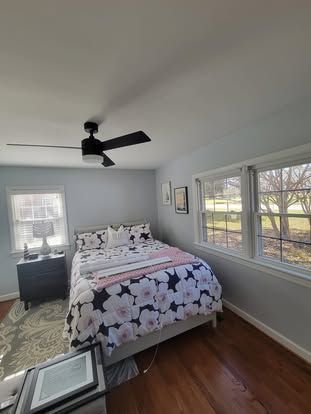 A bedroom with a floral bedspread, a ceiling fan, a bedside table, and two windows looking out onto a grassy landscape.