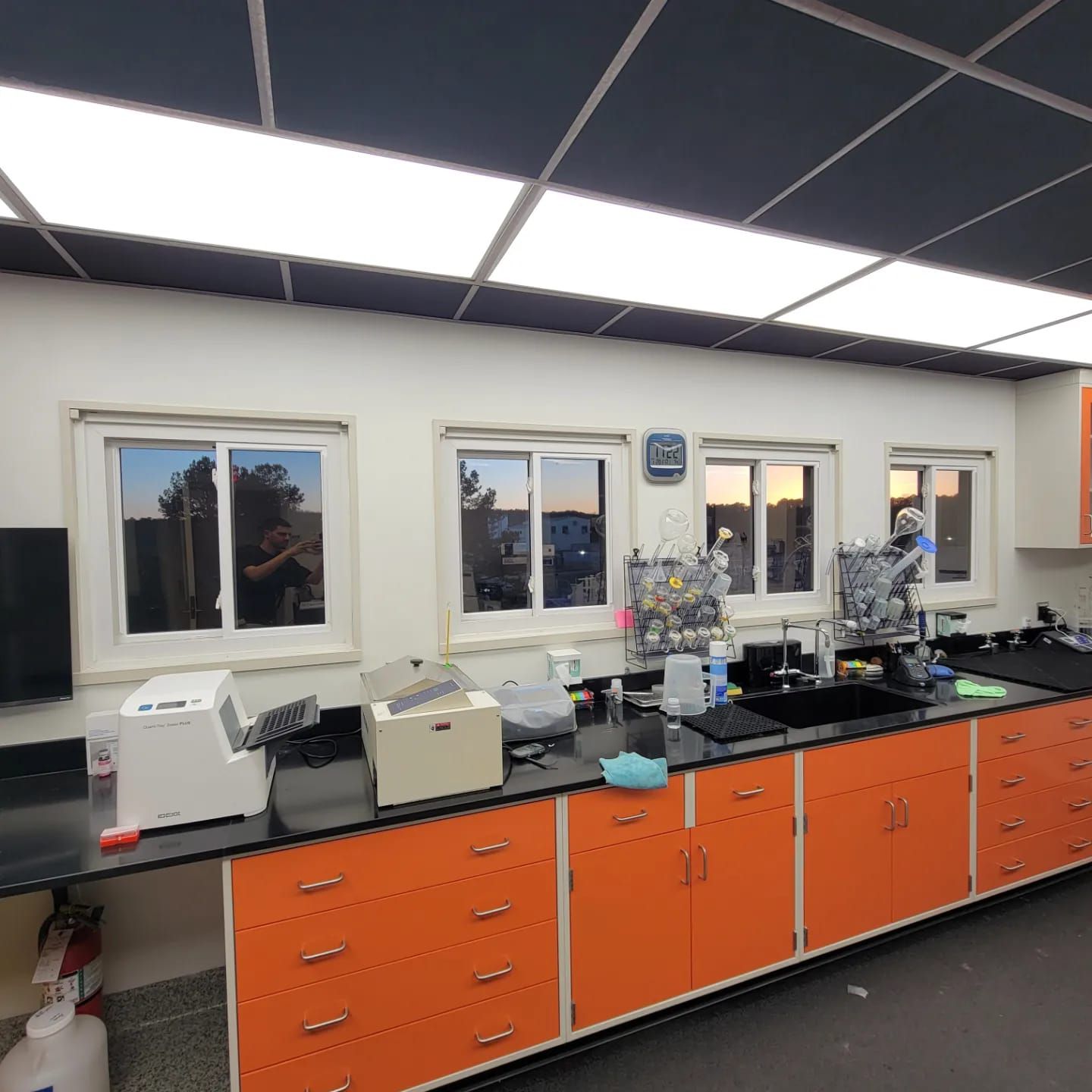 A laboratory workbench with bright orange cabinets and black countertops, featuring windows, equipment, and a sink.
