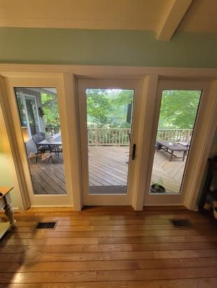 A set of three glass doors opening onto a wooden deck overlooking a leafy backyard with trees and outdoor furniture.