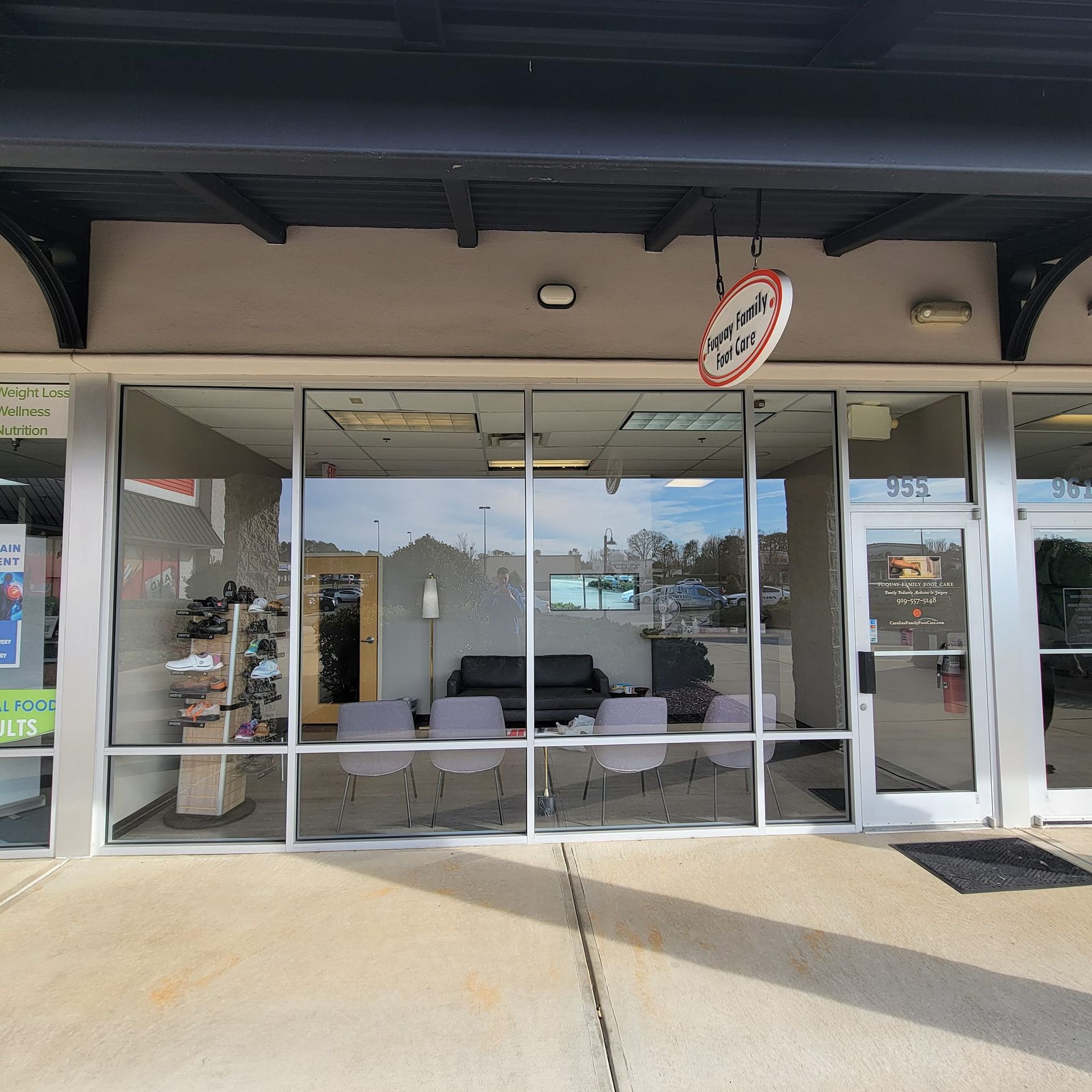 A storefront with large glass windows, a circular hanging sign, a dark couch, and several chairs in the waiting area.