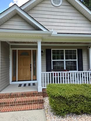 Tan house exterior with a wooden front door, brick steps, a white porch railing, and a green bush in the front yard.